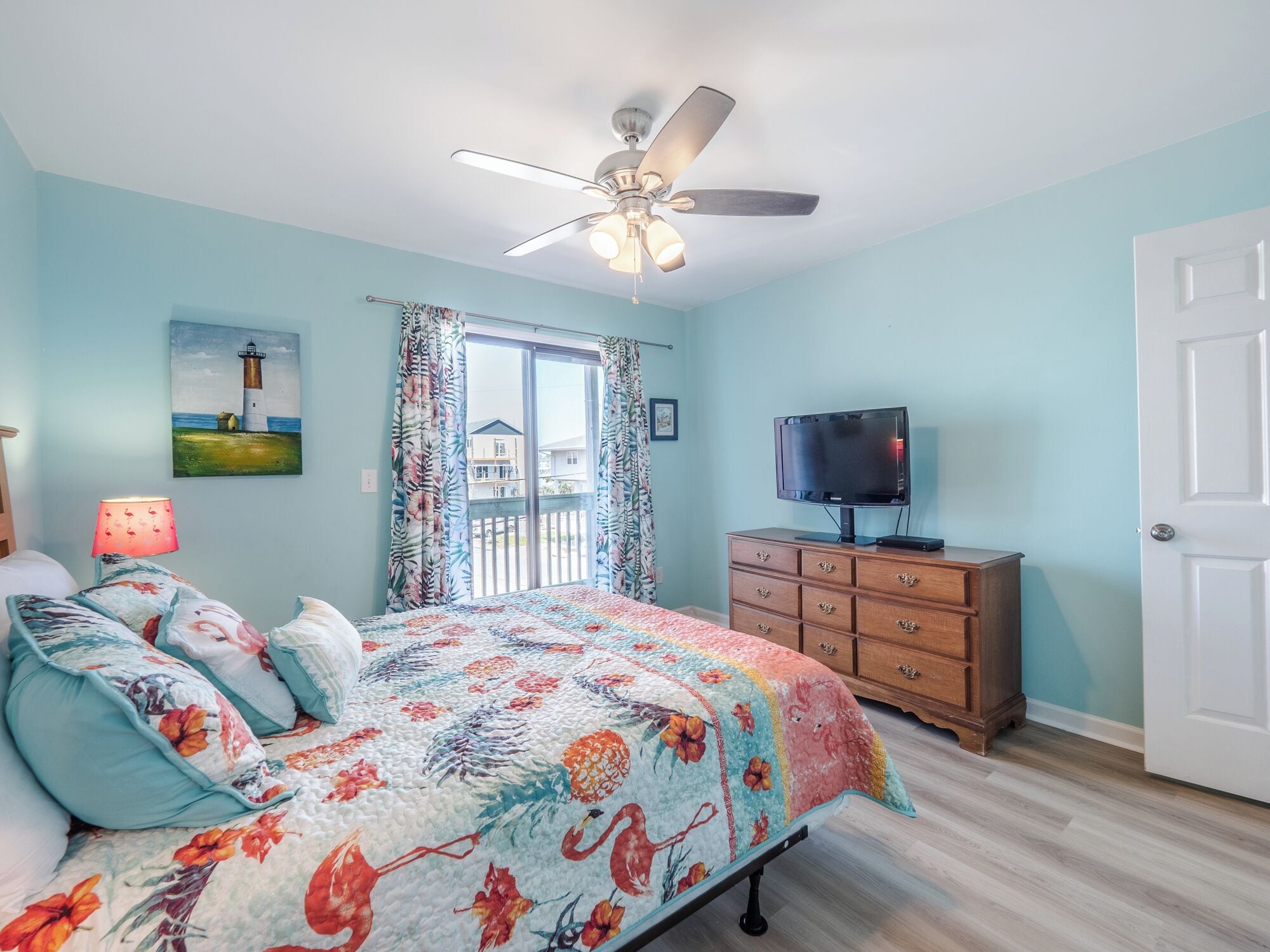A bedroom with a bed, a dresser, a television, a ceiling fan, and a painting of a lighthouse.
