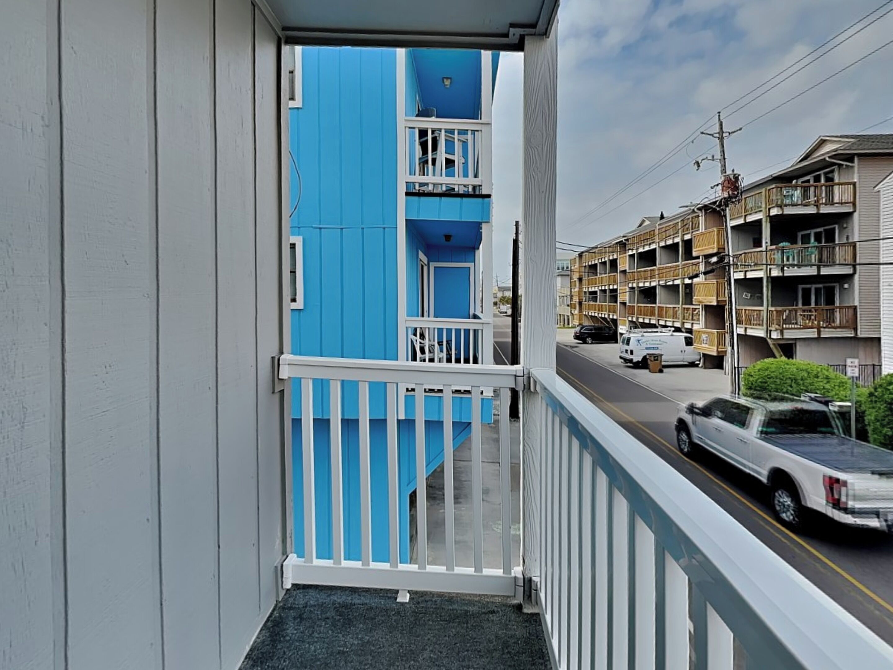 A balcony with a view of a street, a blue building, and a white truck parked on the road.