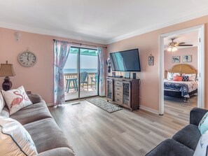 A room with a large window, a flat-screen TV, a sofa, a bed, and a wooden cabinet. - Winds by Carolina Retreats (Carolina Beach)