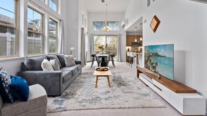A spacious living room with a grey sofa, a coffee table, and a TV. There is a dining area with a table and chairs in the background. - @marbella Lane - Elegant 3BR w/ Marvelous Views (Waianae)