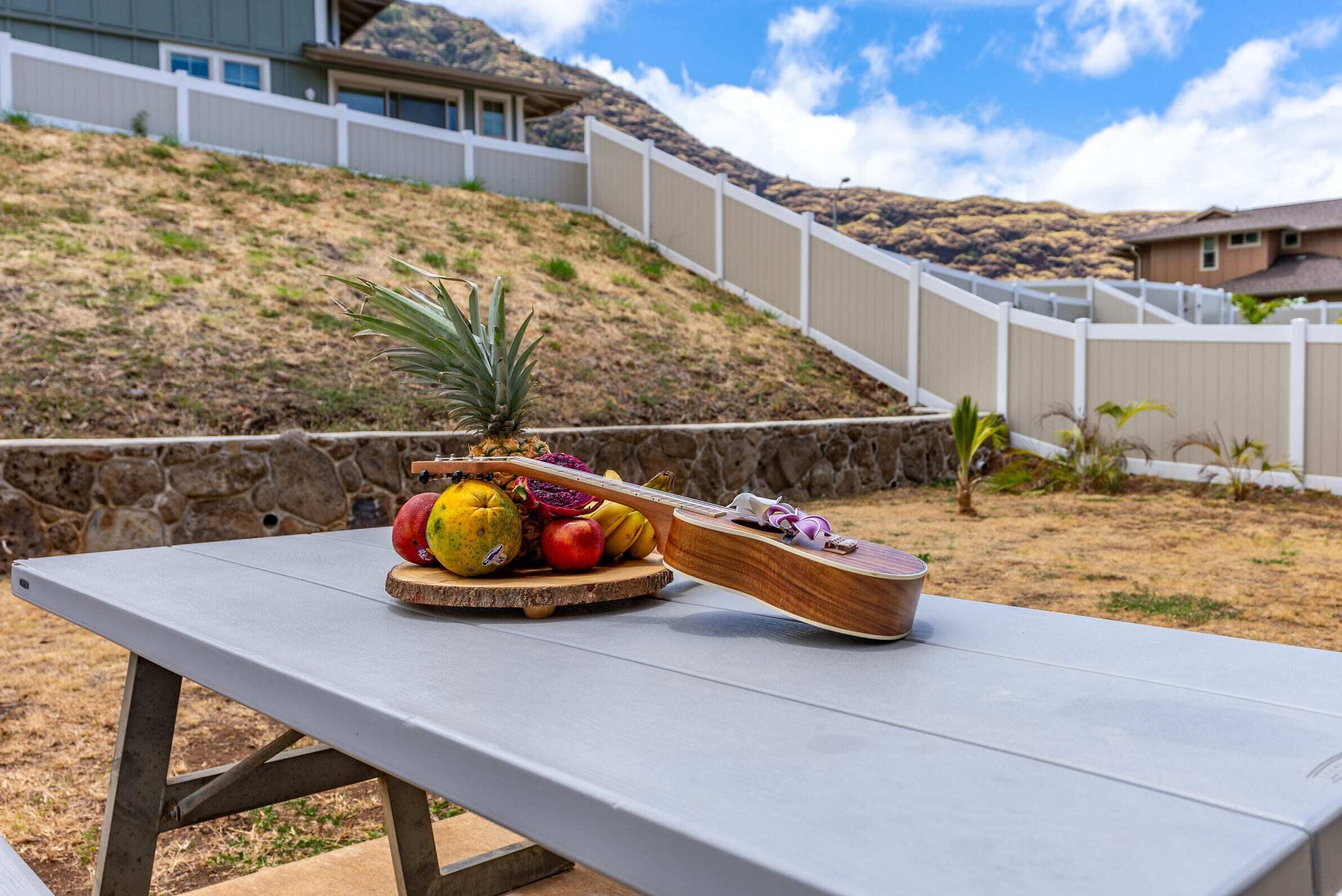 A picnic table with a wooden board holding fruits and a pineapple, set against a hillside with a white fence and houses in the background.