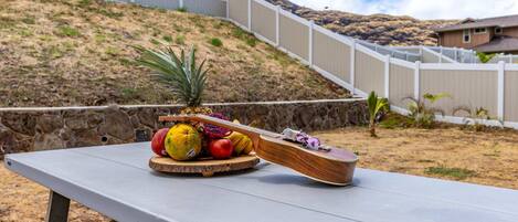 A picnic table with a wooden board holding fruits and a pineapple, set against a hillside with a white fence and houses in the background.