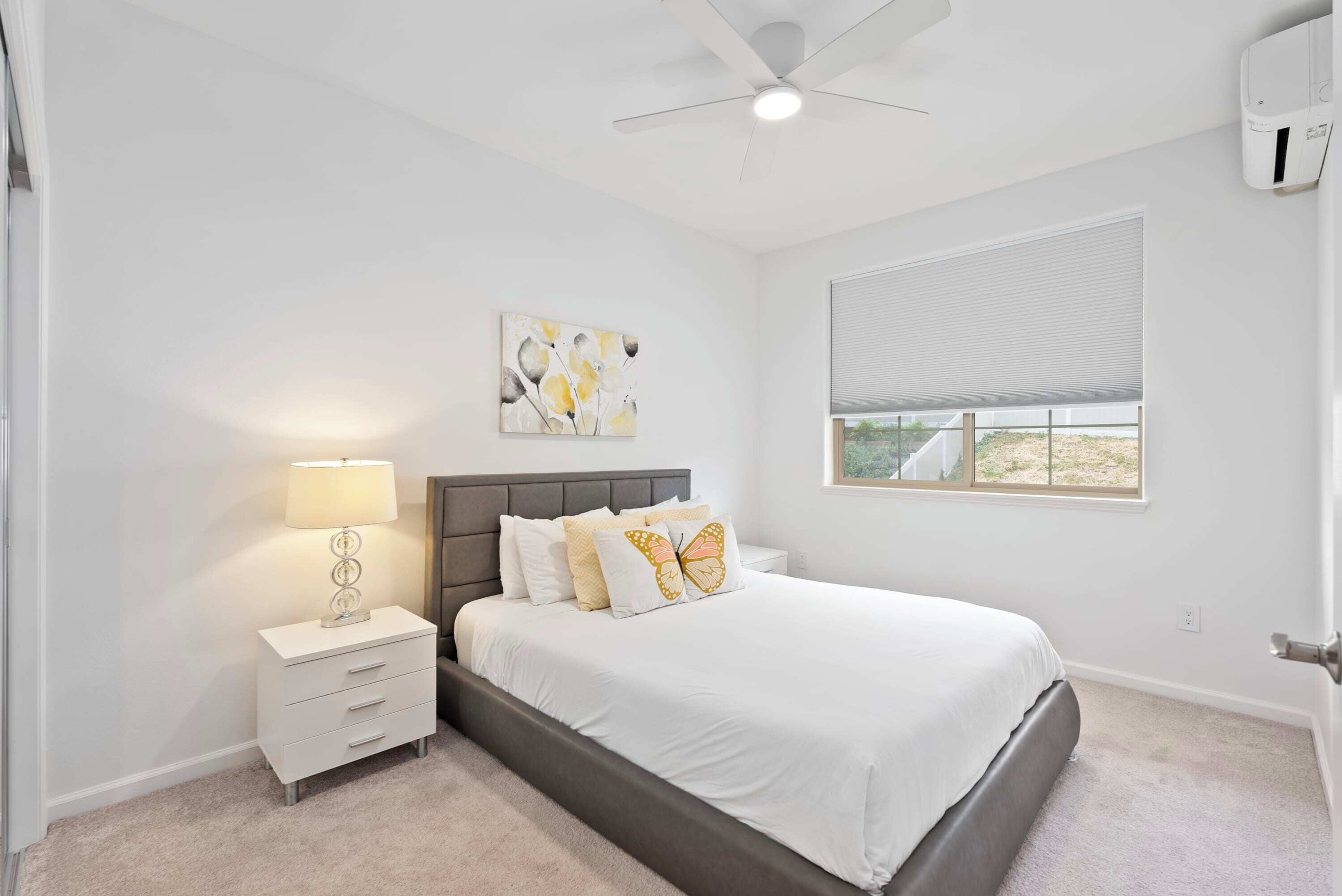 A bedroom with a bed, bedside table, lamp, ceiling fan, window, and artwork.