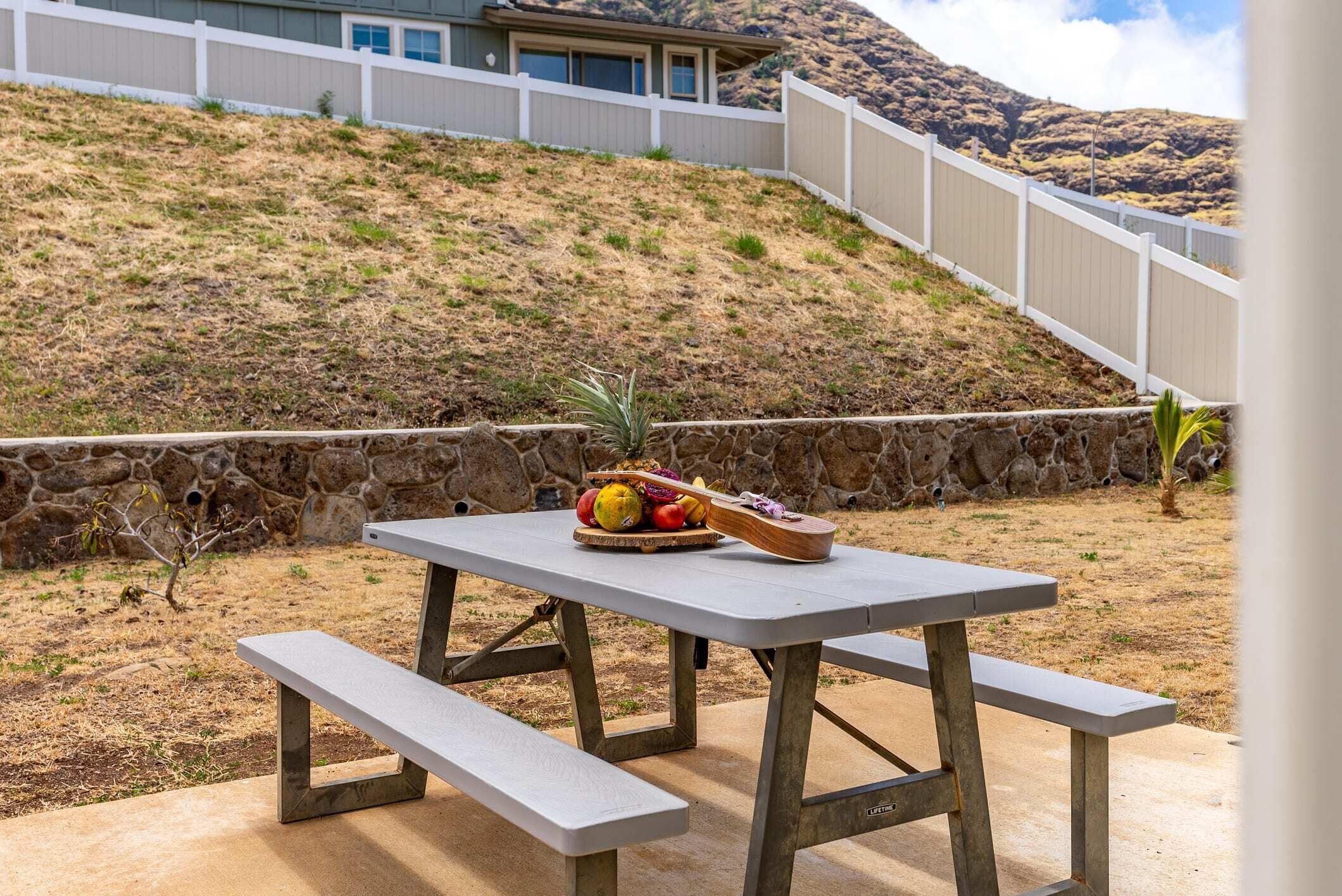 Une table de pique-nique avec des bancs et un panier de fruits, dans un jardin avec un mur de pierres et une clôture en bois.