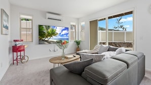 A modern living room with a grey sofa, a coffee table, and a large TV. There is a window with a view of the outdoors. - @ Marbella Lane - Stunning Gem w/ MT & Ocean Views (Waianae)