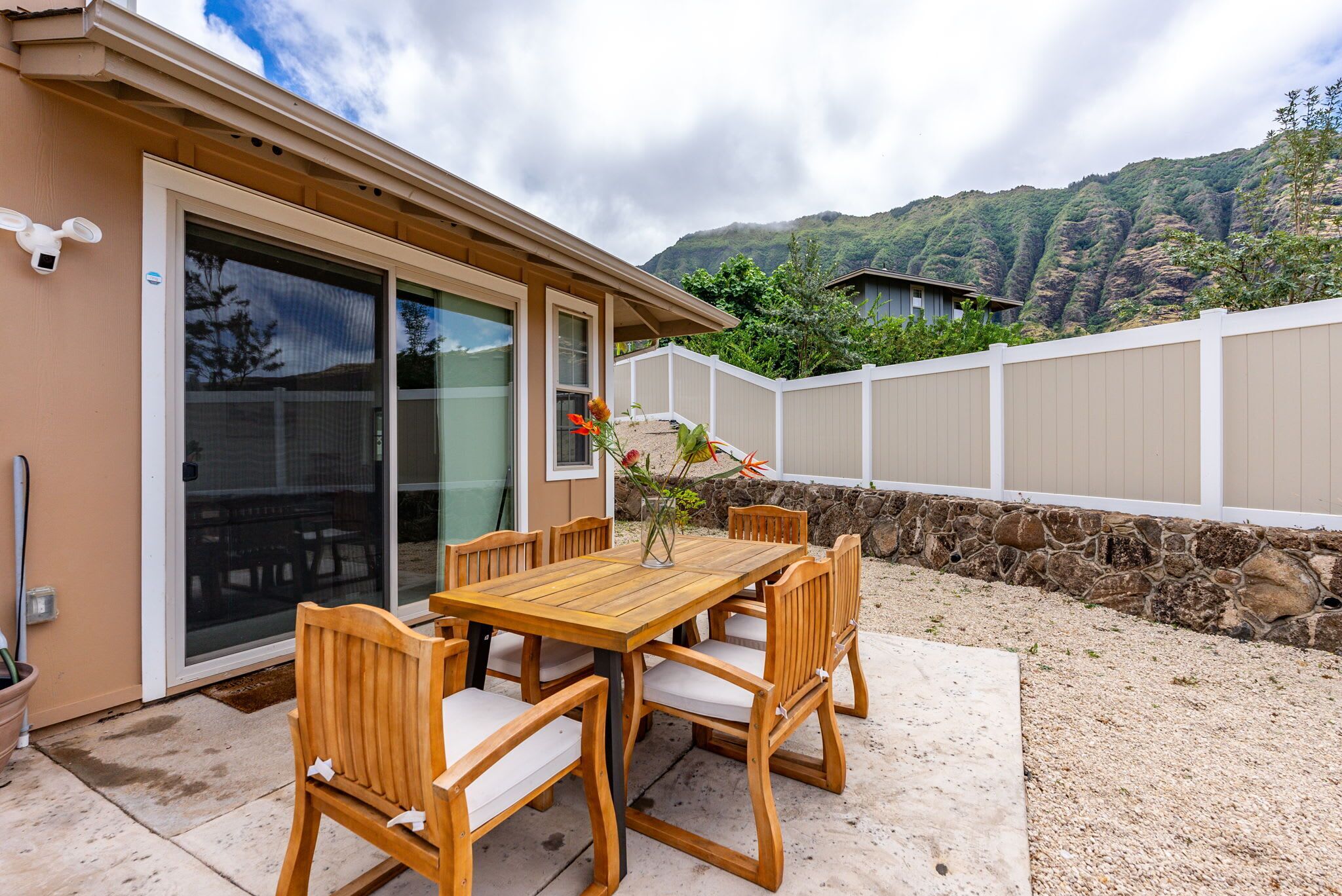 A wooden table and chairs set outside a house with a stone wall and a view of a mountain.