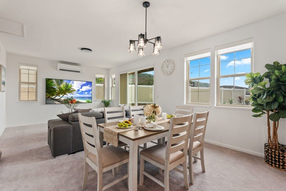 A dining area with a wooden table, beige chairs, and a chandelier. There is a television on the wall and a large window with a view of the outdoors.