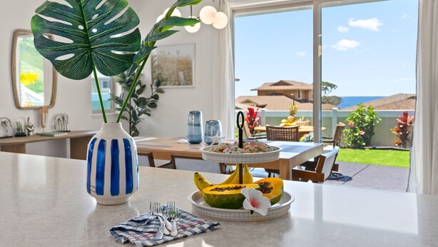 A dining area with a table set for two, a large tropical plant, and a fruit bowl with a banana and a flower. - @ ML Aloha Hoonani 3BR Home MT & Ocean Views! (Waianae)