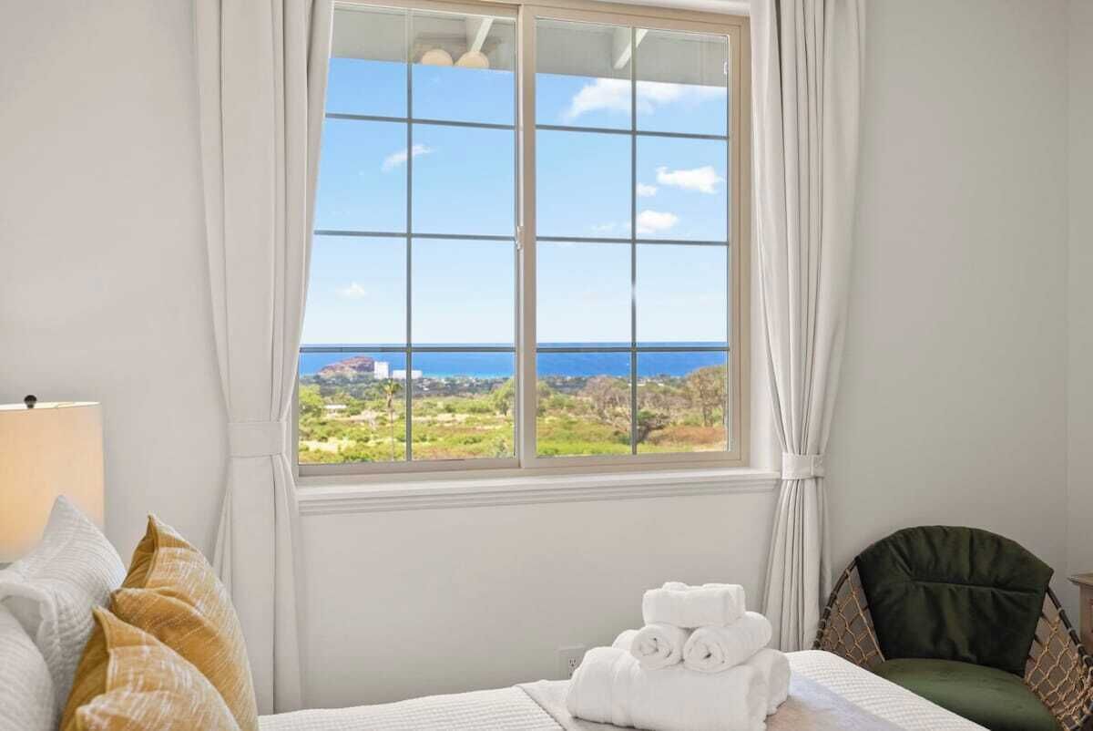 A hotel room with a large window offering a view of the sea and greenery. There is a bed with white linens and a green chair.