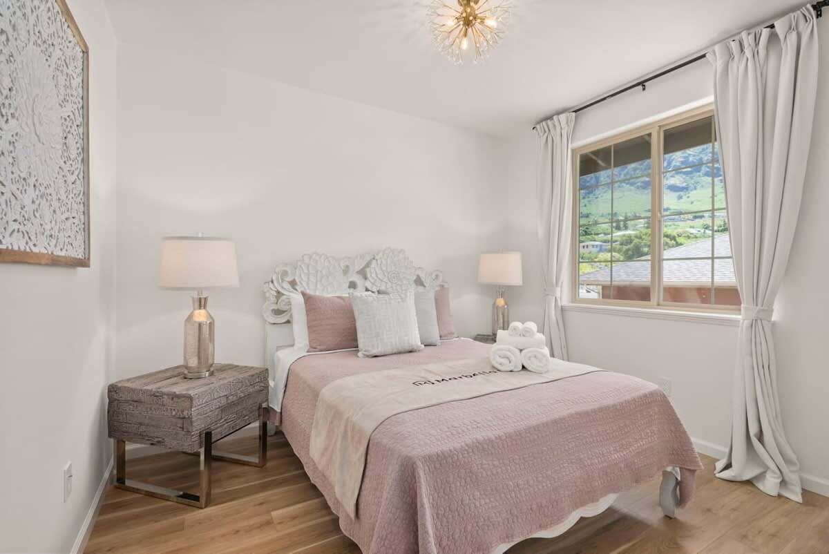 A bedroom with a bed, bedside lamps, a nightstand, a window with curtains, and a view of greenery outside.