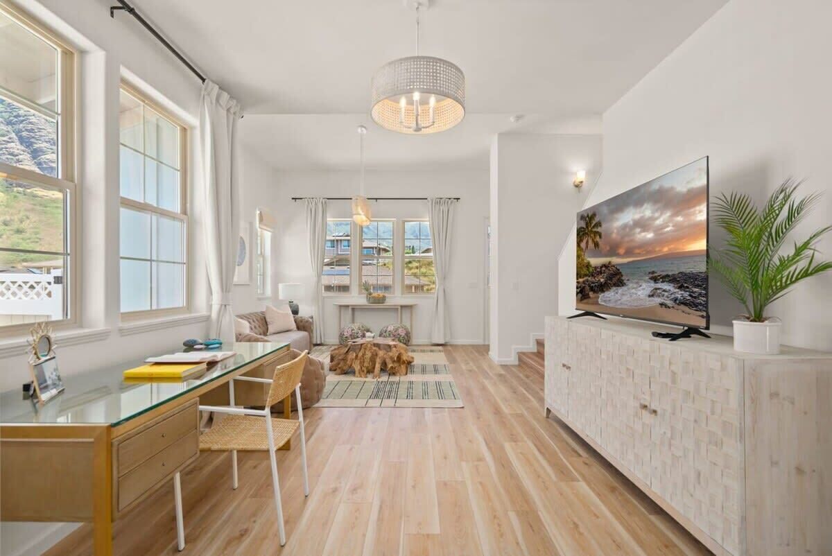 A spacious living room with a large window, a glass desk, a comfortable seating area, and a mounted TV.
