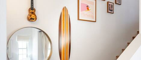 Una escalera con barandilla de madera y una pared decorada con un ukelele, una tabla de surf y fotografías enmarcadas.