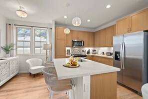 Una cocina moderna con armarios de madera, electrodomésticos de acero inoxidable, una isla central con fruta y sillas blancas.