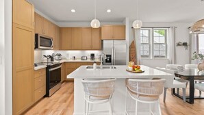 A modern kitchen with wooden cabinets, stainless steel appliances, a central island with two white chairs, and pendant lighting. - @marbella Lane - Paradise Gem W/breathtaking Views (Waianae)