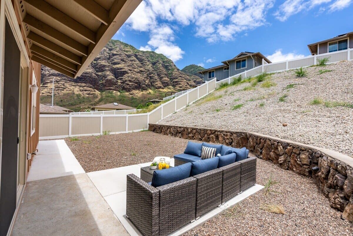 A covered patio with a wicker sofa and a small table, a stone retaining wall, and a view of a mountainous landscape.