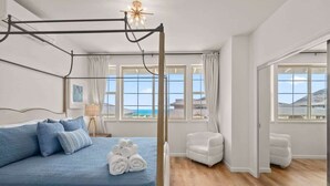 A modern bedroom with a four-poster bed, a white armchair, and a view of the sea through large windows. - @marbella Lane - Paradise Gem W/breathtaking Views (Waianae)