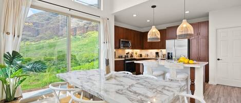 A modern kitchen with a marble island, white chairs, and a view of greenery.