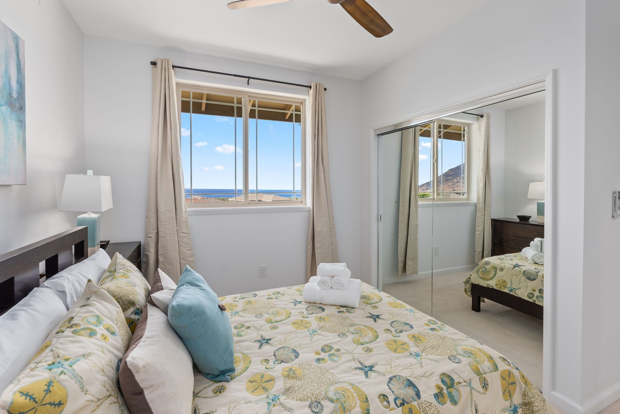 Une chambre à coucher avec un lit, une table de chevet, une lampe, une fenêtre avec des rideaux, un miroir et une vue sur la plage.