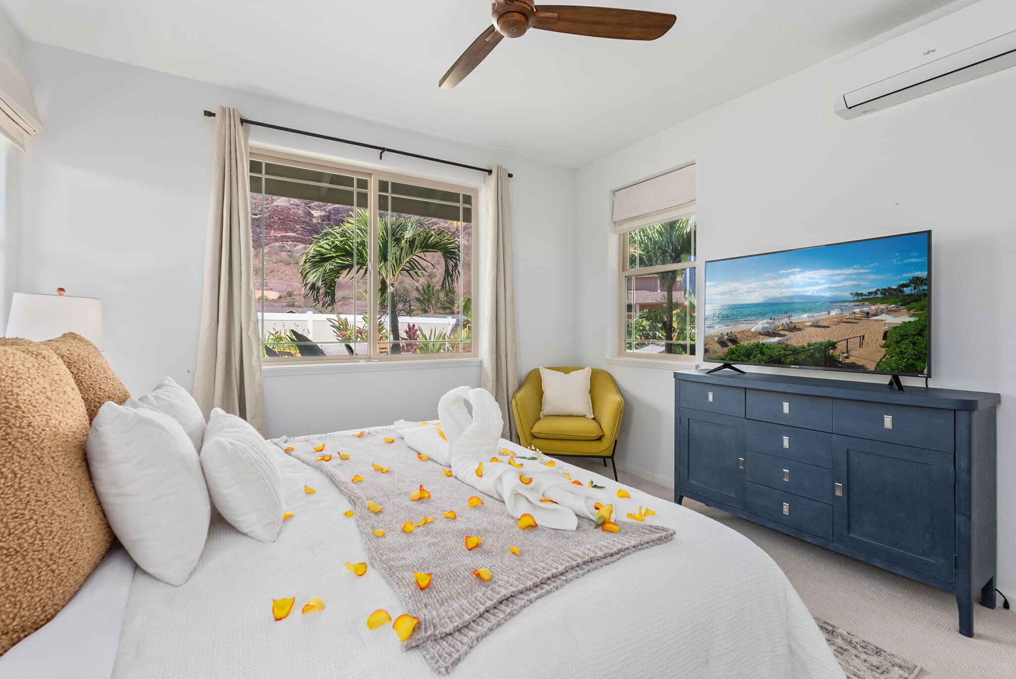 A hotel room with a bed, a television, a ceiling fan, and a view of the beach through the window.