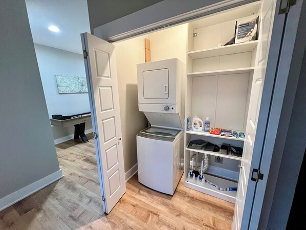 A compact laundry room with a washing machine, a drying rack, and shelves with cleaning supplies. - Beautiful Craftsman Home in Historic Adair Park (Atlanta)