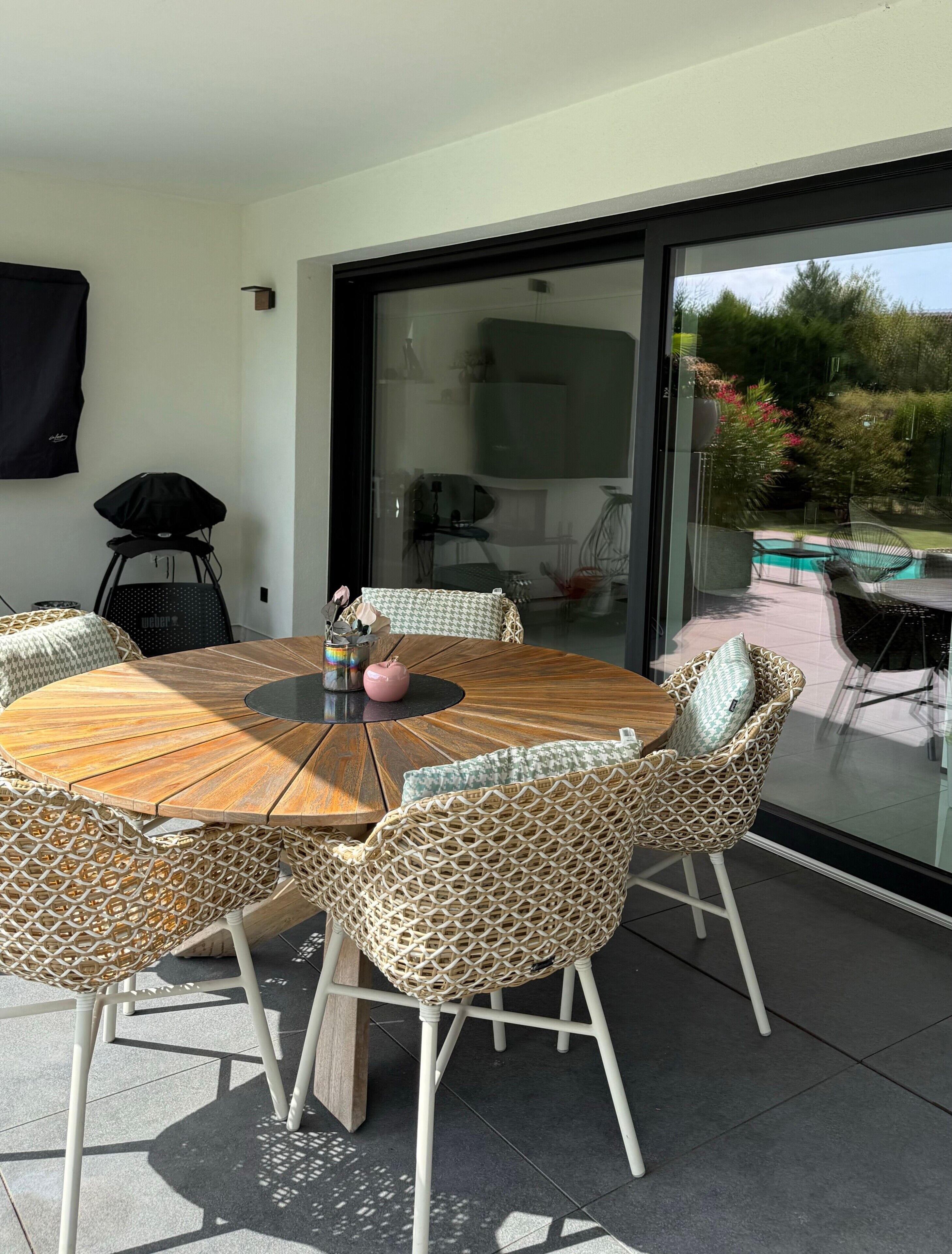 Ein runder Holztisch mit Korbstühlen, eine Glastür zur Terrasse und Blick auf einen Swimmingpool.