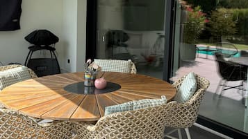 Ein runder Holztisch mit Korbstühlen, eine Glastür zur Terrasse und Blick auf einen Swimmingpool.