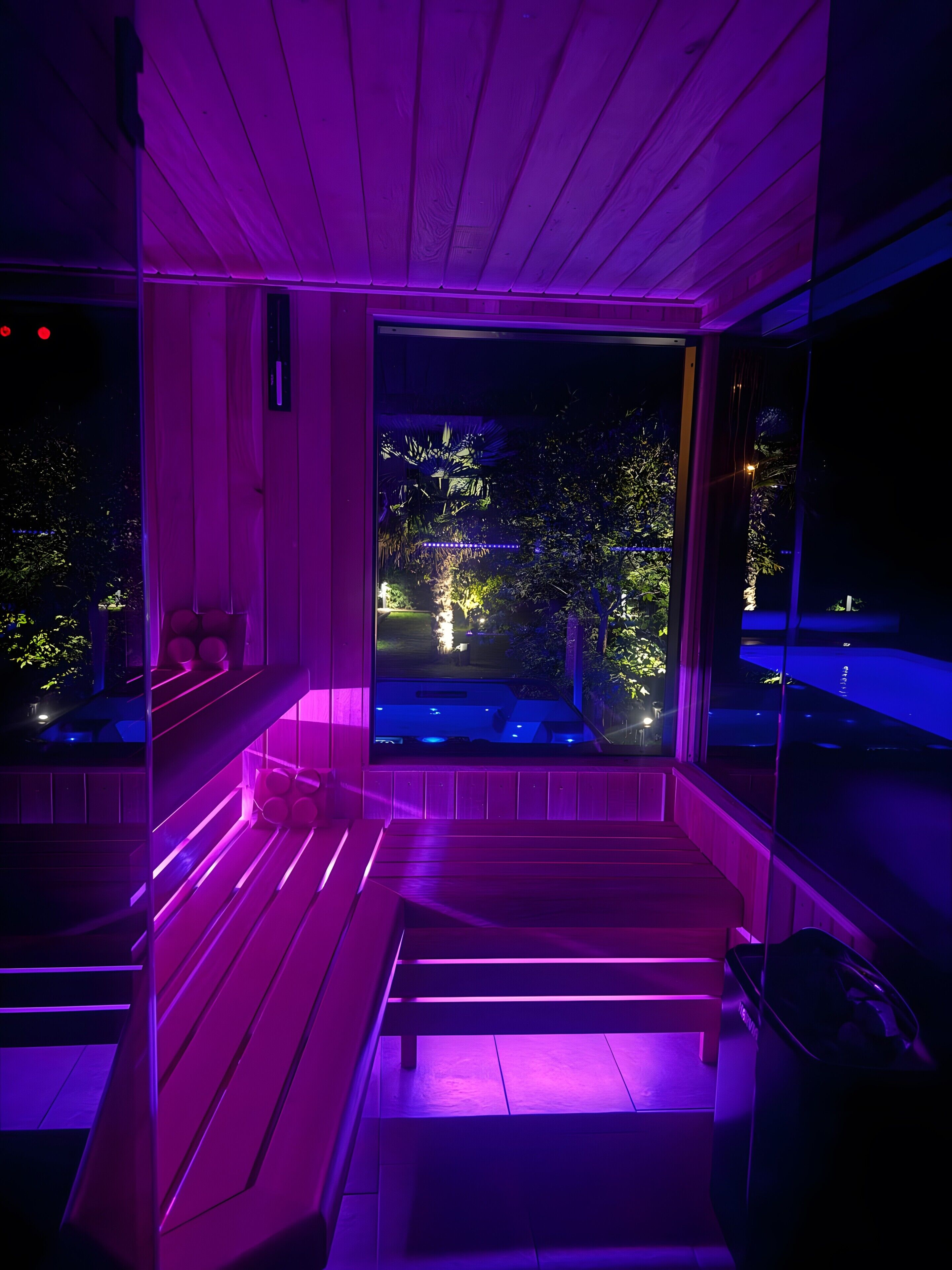 Une pièce avec un banc en bois, une porte vitrée donnant sur l’extérieur et une vue sur un jardin la nuit.