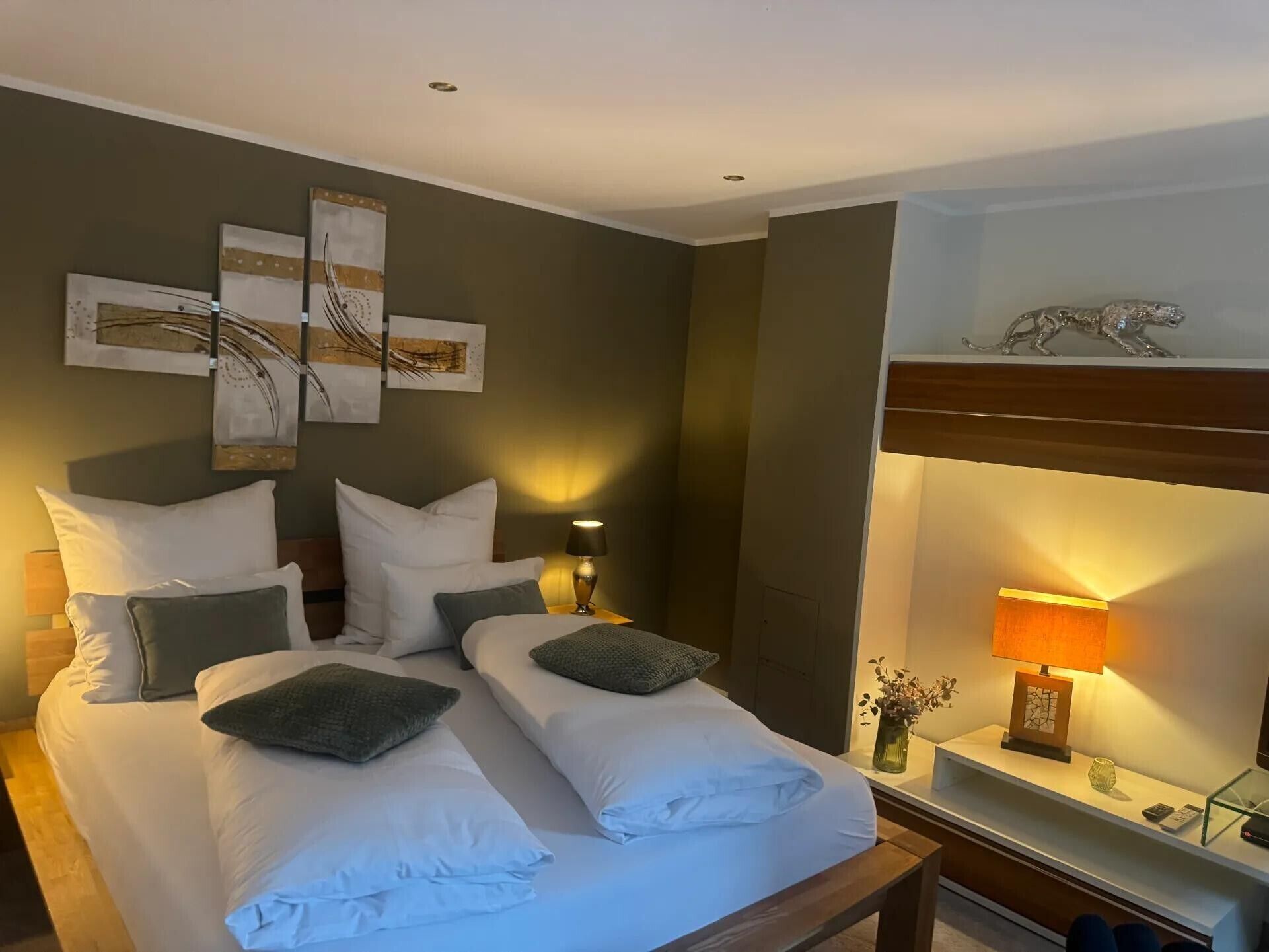 Une chambre d’hôtel avec un lit double, deux tables de chevet, une petite table, une lampe, un élément décoratif mural et une étagère avec un vase et un tableau encadré.