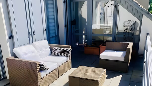 Un espace extérieur aménagé avec des sièges, baigné de lumière naturelle, comprenant un canapé, un fauteuil et une petite table, le tout délimité par un mur blanc et une porte en verre.