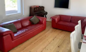Ein Wohnzimmer mit zwei roten Sofas, Holzboden, einem Klavier und einem Fernseher an der Wand.
