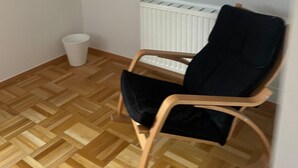 Une pièce d’angle avec un plancher en bois, un radiateur blanc, une chaise noire et une poubelle blanche.