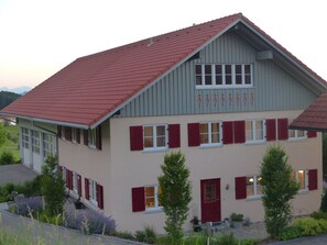 Ein zweistöckiges Gebäude mit roten Ziegeldach, mehreren Fenstern mit Fensterläden und einer Haustür.
