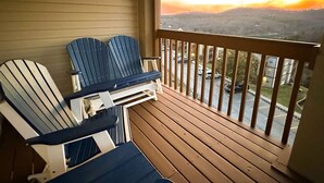 Un balcon avec deux fauteuils Adirondack, une terrasse en bois et une vue sur un parking et des immeubles au coucher du soleil.