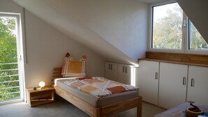A bedroom with a sloped ceiling, a bed with a wooden frame, a nightstand with a lamp, and a window with a view of trees. - Single Room with Wi-Fi (Wäschenbeuren)