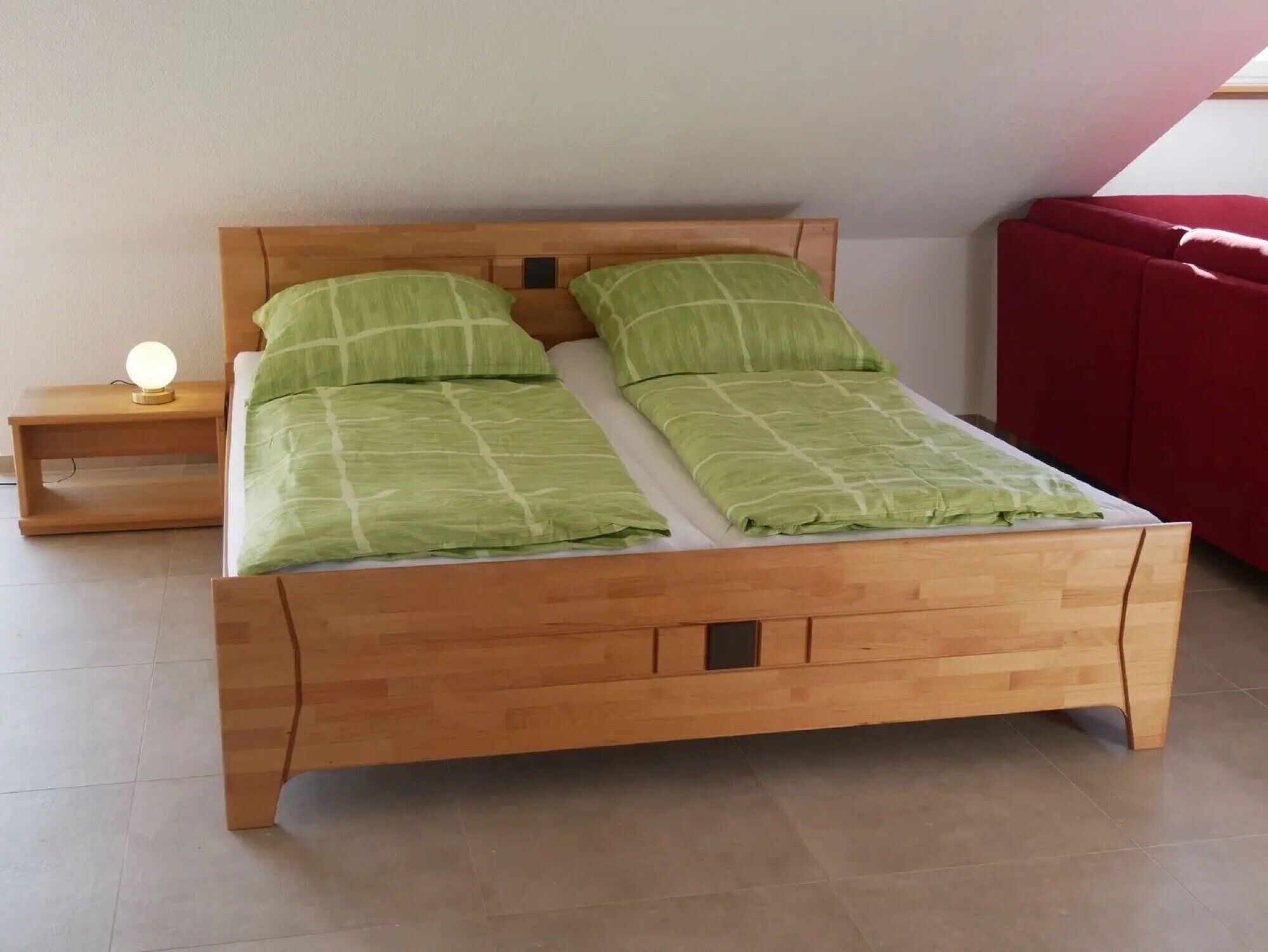 Un lit en bois avec une tête de lit, deux oreillers, une table de chevet avec une lampe, et un canapé rouge en arrière-plan.