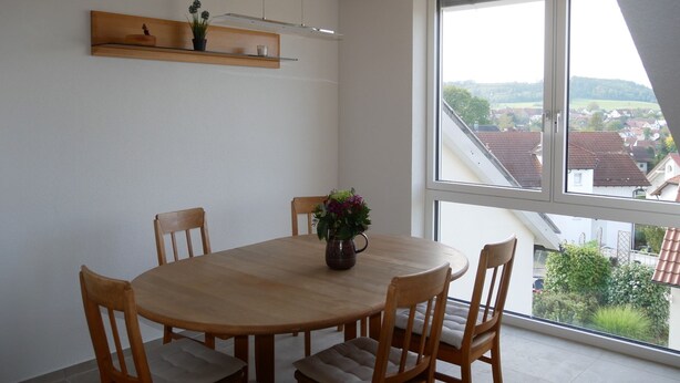 A dining area with a round wooden table, four chairs, a vase with flowers, a wooden shelf above the table, and a large window with a view of buildings and greenery.
