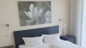 Una cama bien tendida con sábanas blancas, un cabecero oscuro y una obra de arte en la pared con una flor.
