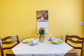 Una mesa de comedor con mantel blanco, dos sillas de madera, un calendario, un florero con flores y una lata de cerveza.
