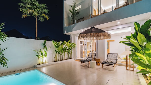 A modern outdoor pool area with a lounge chair and umbrella, surrounded by lush greenery and a two-story building. - Villa Balinay (Canggu)