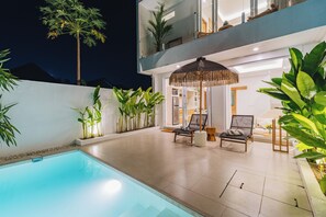 A modern outdoor pool area with a lounge chair and umbrella, surrounded by lush greenery and a two-story building. - Villa Balinay (Canggu)