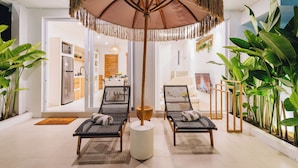 A modern outdoor seating area with two wicker chairs and a central round table, surrounded by lush green plants and a large woven umbrella. - Villa Balinay (Canggu)