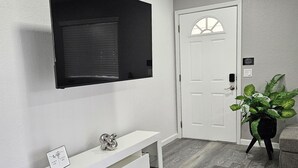 A modern living room with a flat-screen TV mounted on the wall, a white console table with a decorative item, a potted plant, and a closed door.