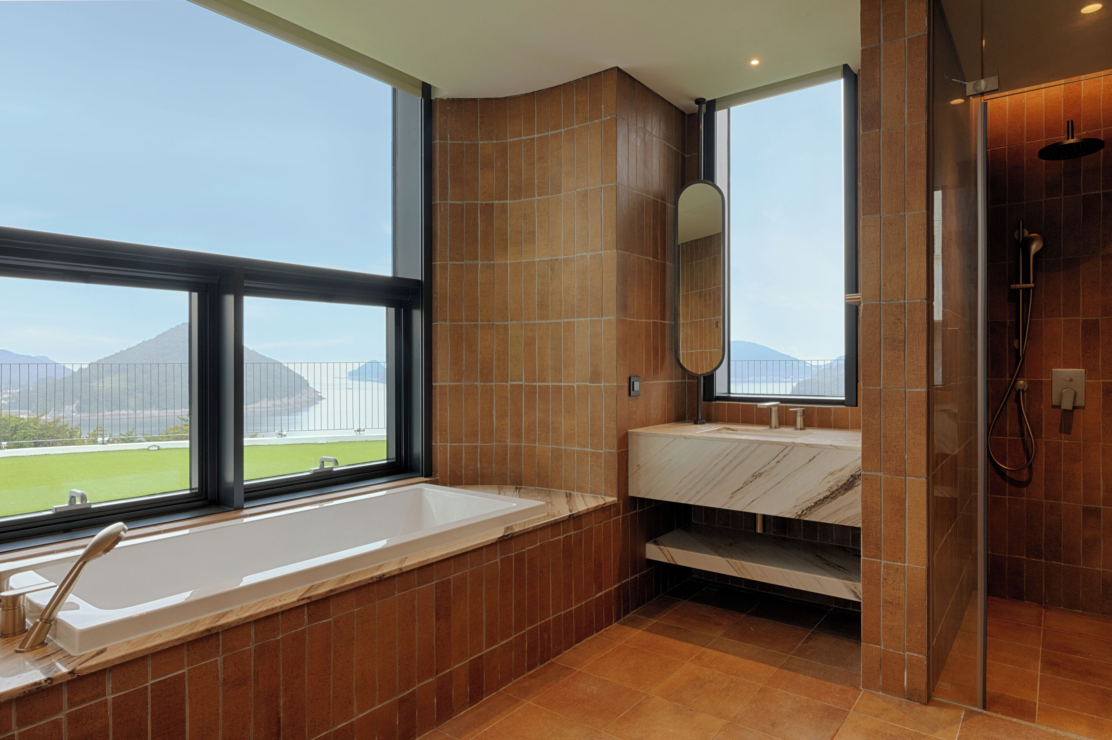 A modern bathroom with a large bathtub, a sink, and a shower. There is a large window providing a view of a mountain and a field.