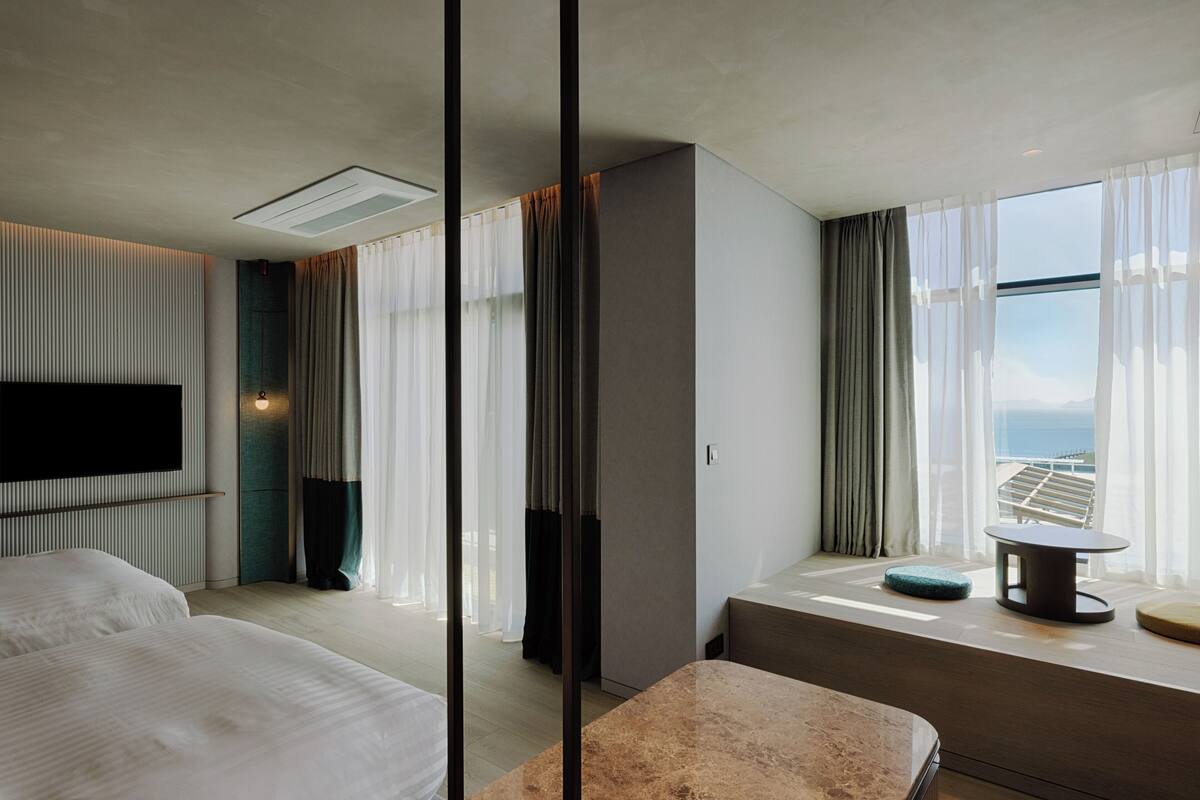 A modern hotel room with two beds, a large window with curtains, a flat-screen TV, and a small table by the window.