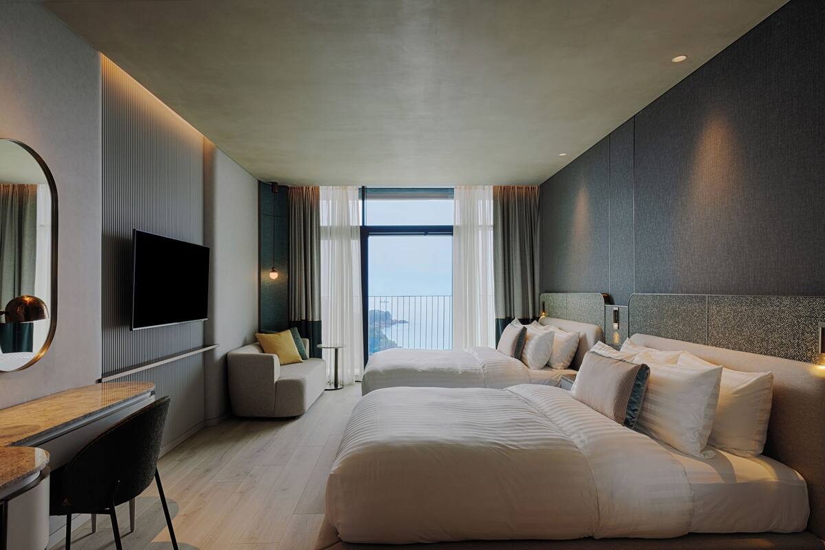 A modern hotel room with a large bed, a desk with a chair, a flat-screen TV, and a view of the sea through the window.