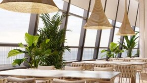 Un espace de restauration moderne, doté de hauts plafonds, de grandes fenêtres et de luminaires suspendus. Les tables et les chaises sont disposées pour les repas.