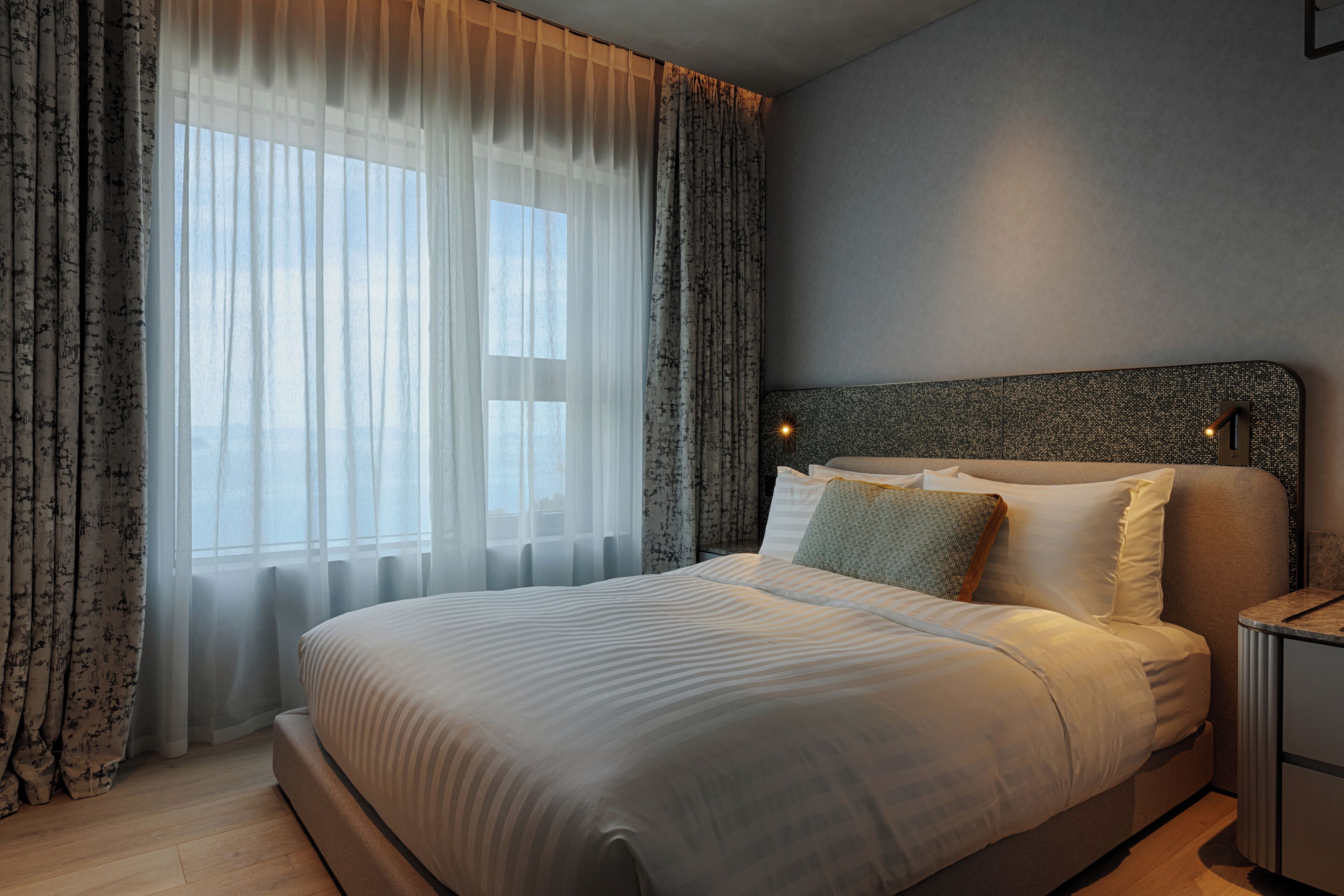 Moderni hotellihuone, jossa on suuri sänky, kuvioitu pääty ja läpikuultavat verhot ikkunoissa.