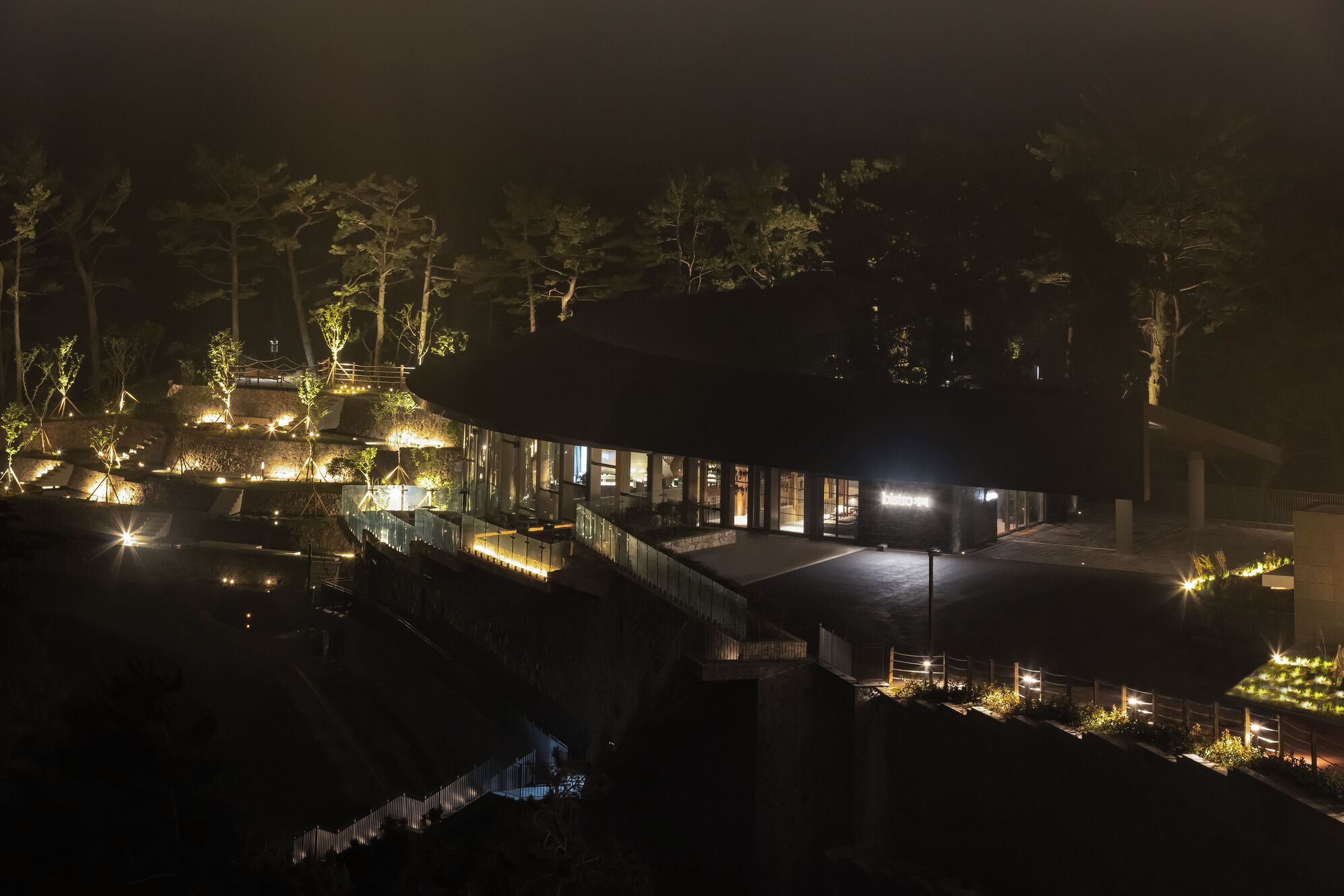 A modern building with a glass facade, surrounded by trees and illuminated pathways.