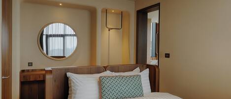 Ein Hotelzimmer mit einem Bett, einem Nachttisch, einem Spiegel und einem runden Fenster.
