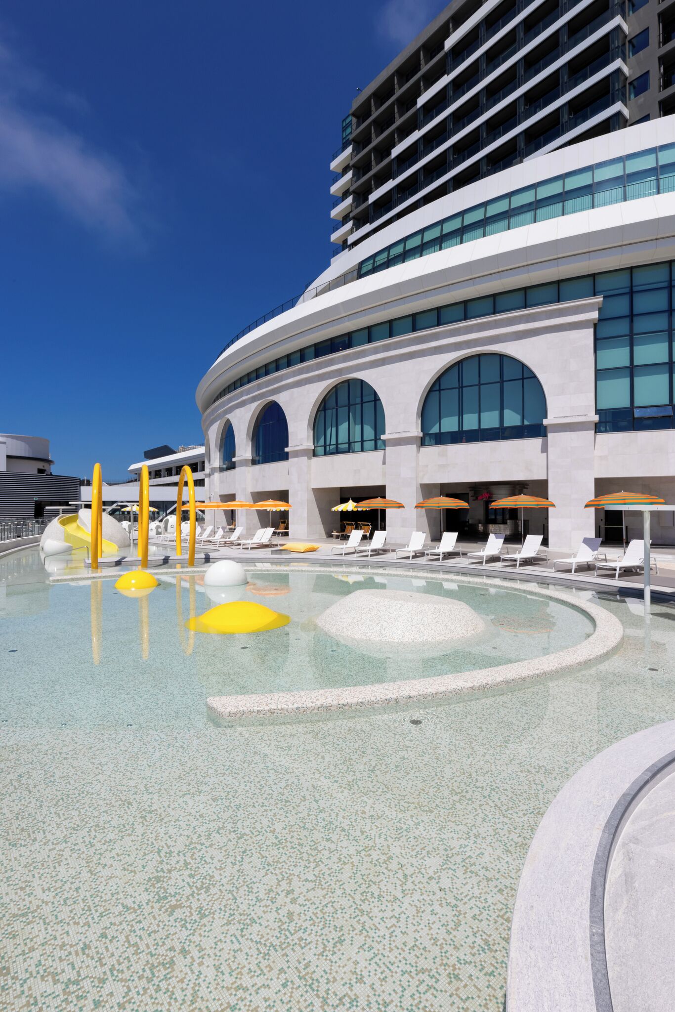 A modern hotel with a pool area featuring sun loungers and umbrellas, a large building with arched windows, and a clear blue sky.